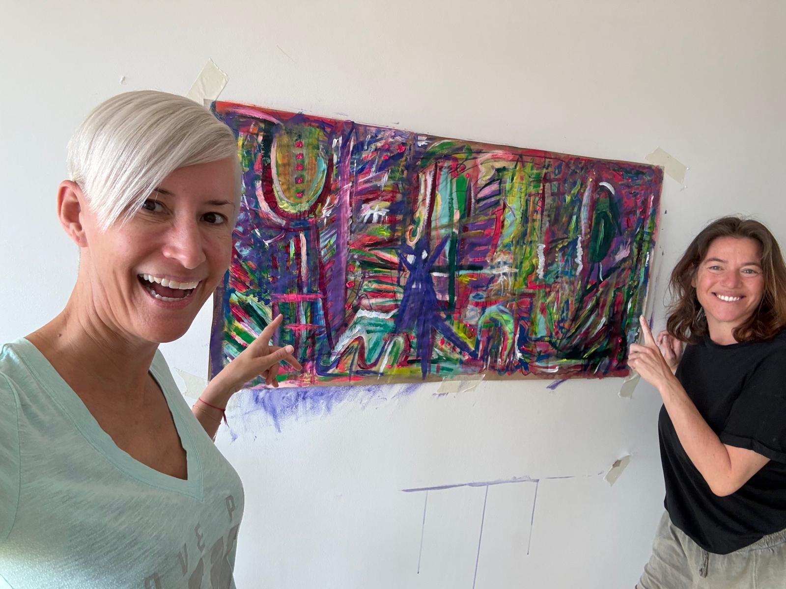 Two participants smiling and pointing at a colourful abstract painting taped to the studio wall